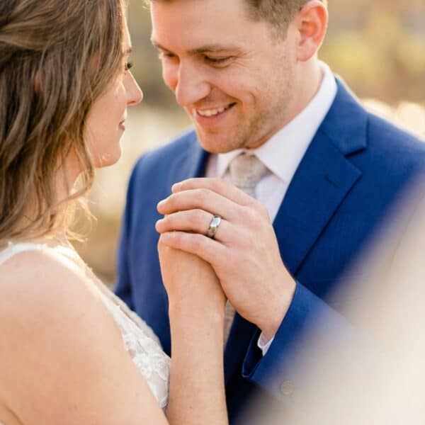 The wedding couple stand face to face smiling at each other and holding one another's hand. The man is wearing a classically styled mens wedding band - a domed polished silver ring that is made with tungsten.