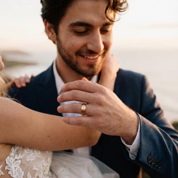 A man smiles happily as he carries his bride on the beach. Wearing a smart suit, the man is wearing his gold tungsten wedding ring.