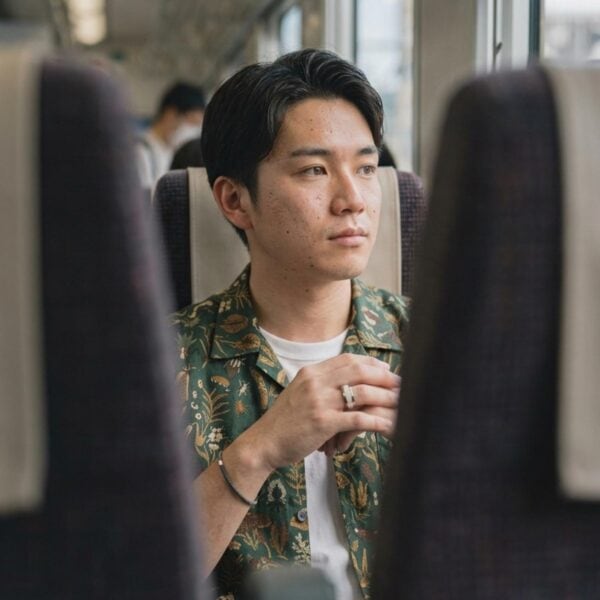 A man sits on a train looking out of the window. He holds up one of his hands in front of him. He wears a silver tungsten ring that has a mother of pearl inlay.