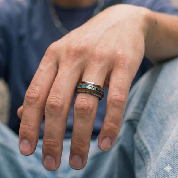 A close up of a mans handing wearing a striking ring. It is a silver tungsten ring that has inlays of natural wood and abalone shell.