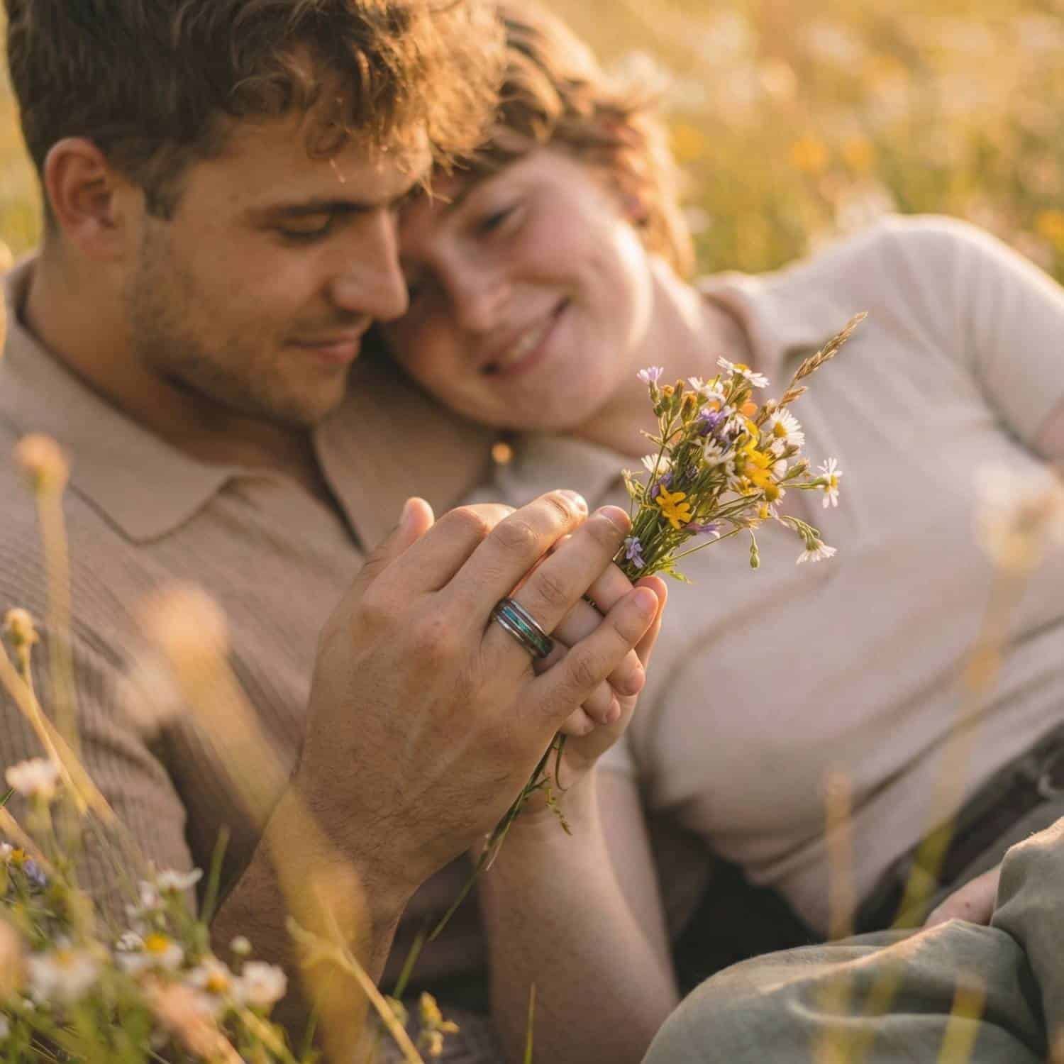 A blissful scene of a happy couple relaxing on the long grass in the countryside. Smiling and leaning against each other, the man holds a small bunch of yellow flowers. On his finger, he wears a silver tungsten ring that features inlays of natural wood, and abalone shell.