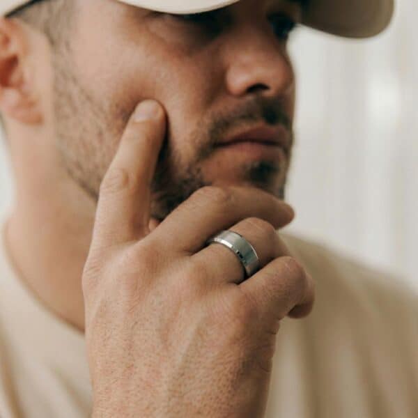 A close up of a mans face who is wearing a matching cream t-shirt and cap. He has his hand held up to his face. He is wearing a silver tungsten ring. It has polished bevelled edges and a brushed centre section.