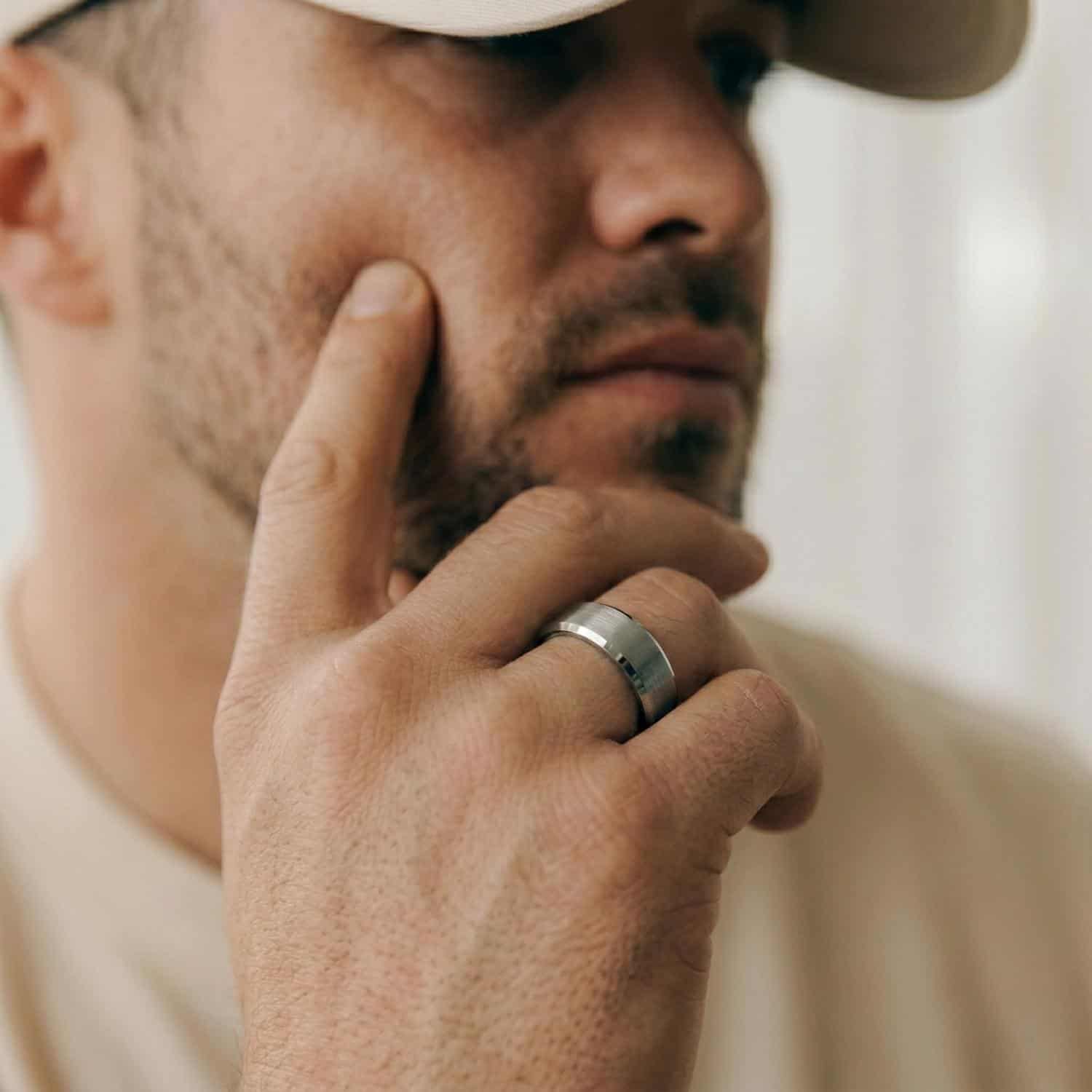 A close up of a mans face who is wearing a matching cream t-shirt and cap. He has his hand held up to his face. He is wearing a silver tungsten ring. It has polished bevelled edges and a brushed centre section.