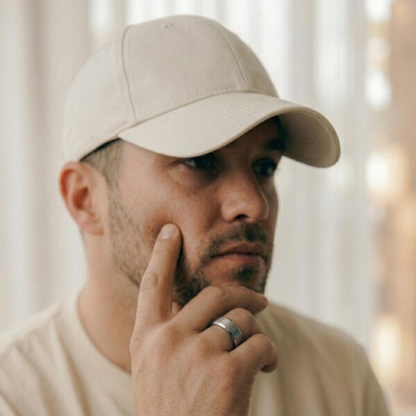 A man looks lost in thought, wearing a cream cap and a matching cream t-shirt. He has his hand held up to his chin. He is wearing a silver tungsten ring that has a brushed centre and polished bevelled edges.