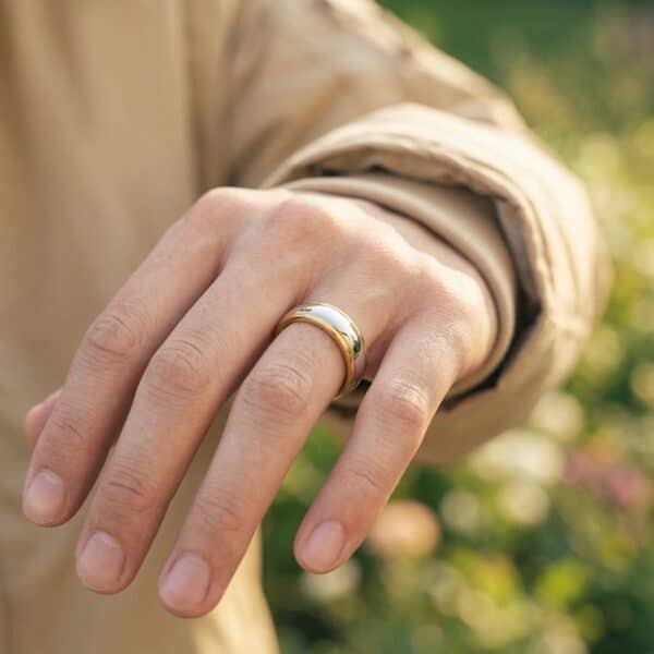 A close up image of a mans hand. He is holding his hand up to the camera to show his ring. It is a gold and silver ring made with tungsten. It has a polished finish. It looks likely to be a mans wedding ring.