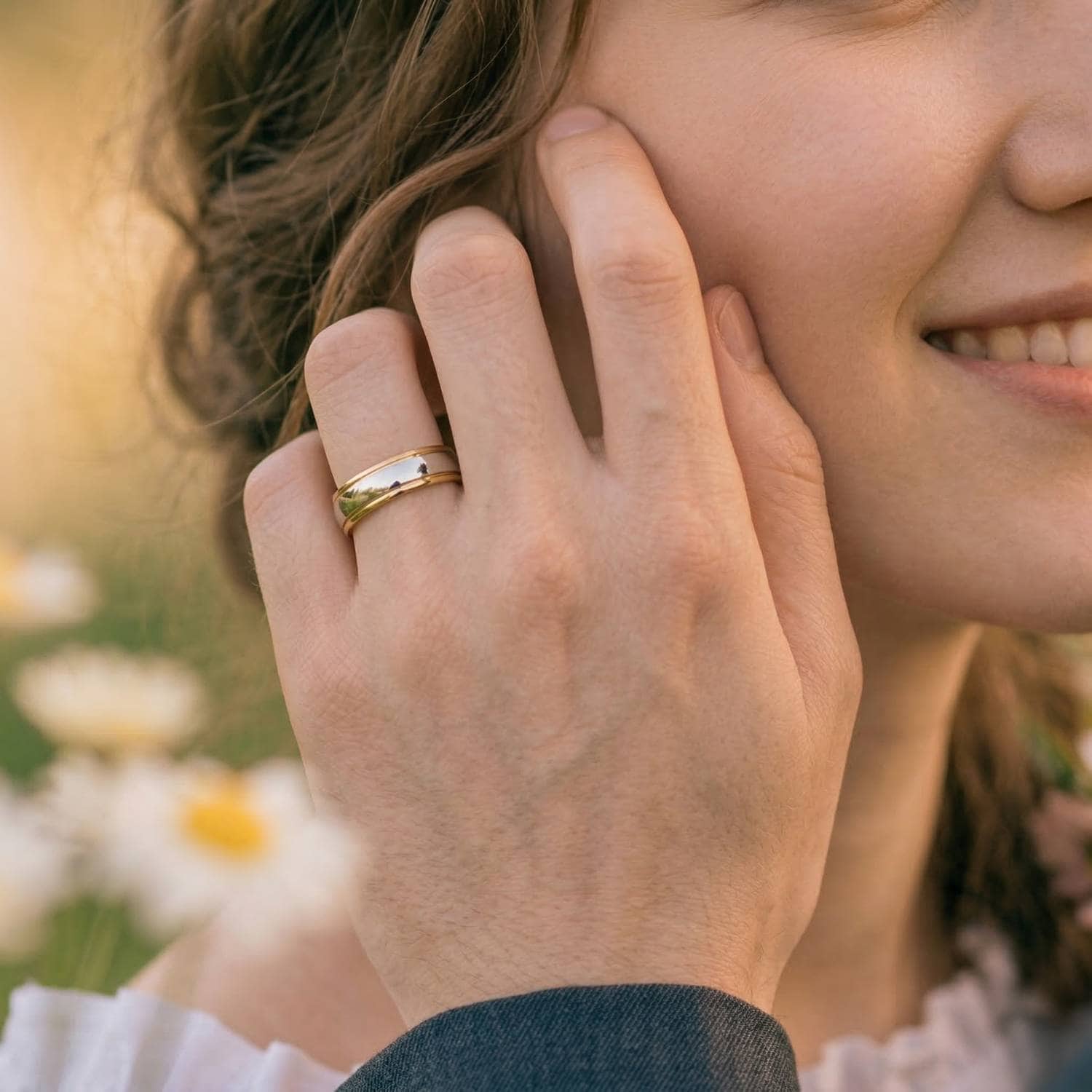 With white and yellow daisies behind them, the photo shows a close up image of a brides face. Her man has his hand held to her face in an affectionate manner. The man is wearing his new wedding band. It is a two tone tungsten ring of silver and gold and has a polished finish.