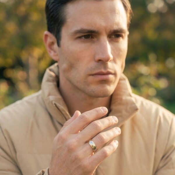 A man looks slightly to one side of the camera lens. Wearing a warm coat, and with bushes in the background, a man has his hand raised up in front of his chest. He is wearing a shiny silver and gold tungsten ring.