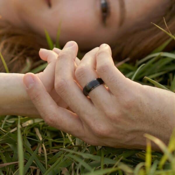 A couple lay on the grass holding hands. You can see a small part of the face of the woman. The couple are holding hands. The man is wearing a black tungsten ring. The centre of the ring is raised and has a brushed finish.