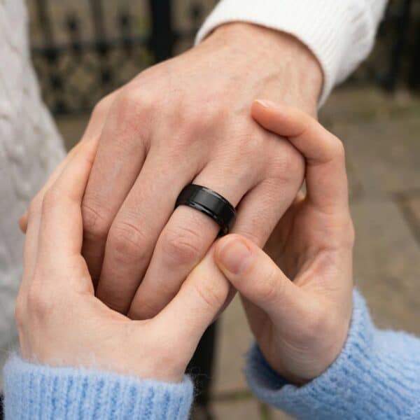 A man puts his hand forward to show his black ring. He is wearing a black tungsten ring that has a raised brushed centre.