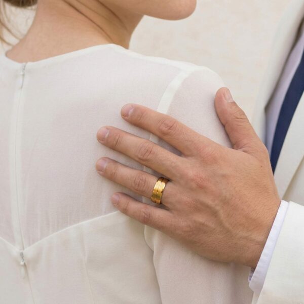 A newly married couple stand close together. The man rests his hand on his partners shoulder. He is wearing his new wedding band which is a gold titanium ring featuring a centre groove and bevelled edges.