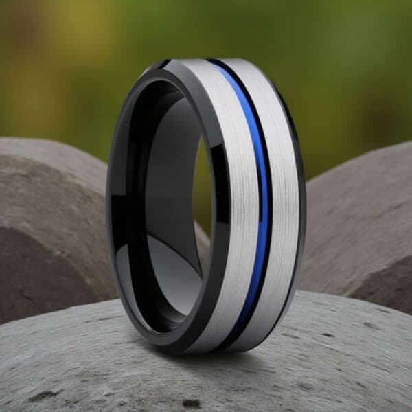 A close up view of a ring that is standing upright on some grey stones. The ring has two raised silver sections which are separated with a blue groove. The edges are bevelled and are polished black.