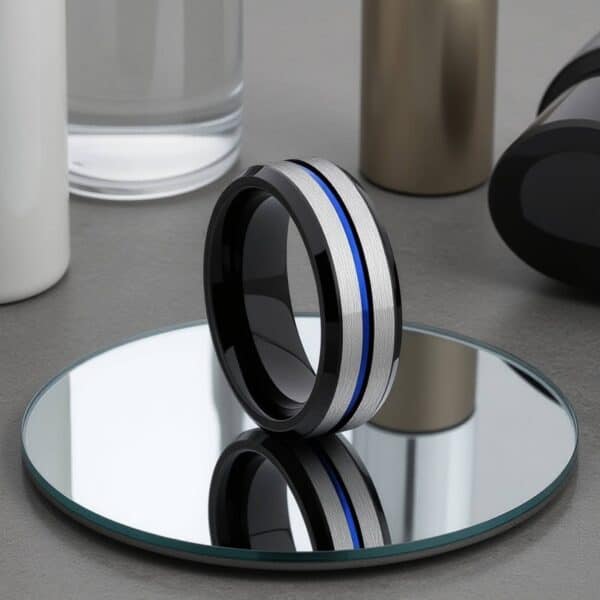 A stylish tungsten ring is standing upright on a circular mirror. The ring has a brushed silver section with a blue groove that runs through the centre of the ring. The edges and inside of the ring are polished black.