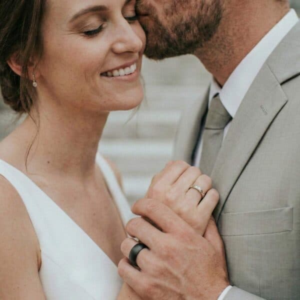 Kissing the bride, her face breaks into a wide smile. Holding hands together, the couple stand close together. On display are their new wedding rings. While the woman is wearing a silver band, the man has a black wedding ring made with tungsten. It has a slightly brushed finish.