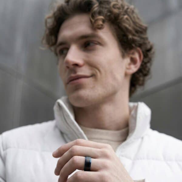 Wearing a white fleece and looking to one side, a man is smiling slightly. He has his hand held out in front of him. He is wearing a black tungsten ring that has blue edges.