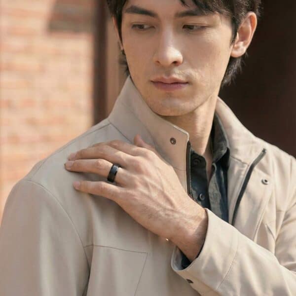 Wearing a raincoat, the man reaches his hand up to his shoulder. He wears a ring on his wedding finger. It is a domed black tungsten ring that has silver edges.