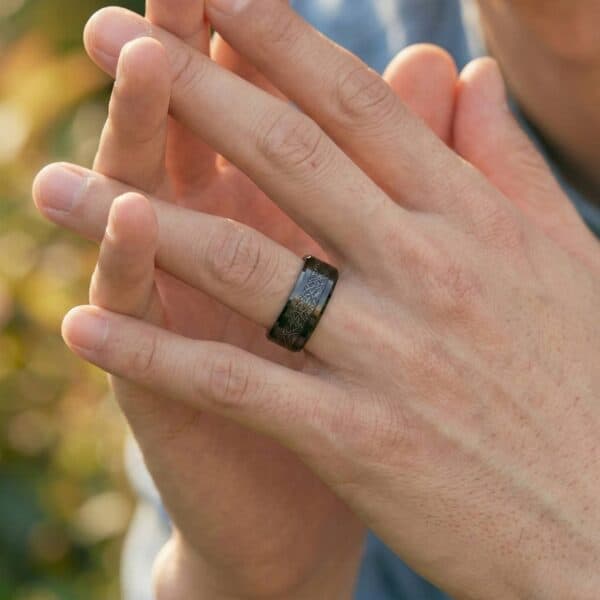 A close up of a pair of hands clasped together. On the mans hand is a black tungsten ring that has a centre section of faux meteorite.