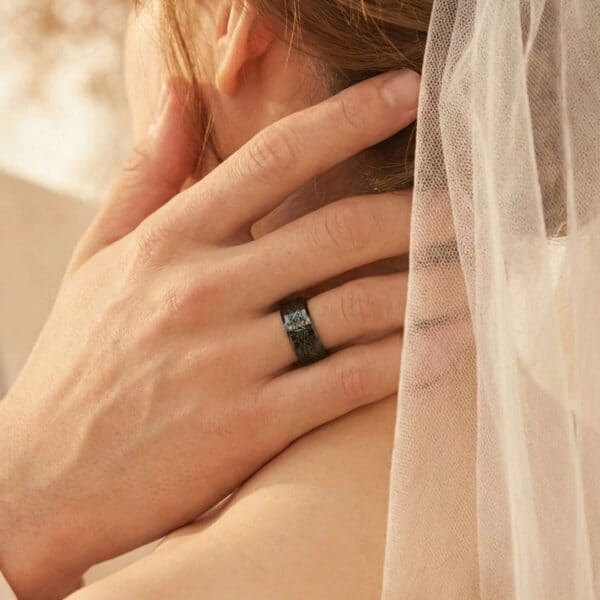 During the wedding ceremony, the couple come together for a kiss. The man takes his bride's face in his hands. He is wearing a black tungsten ring that features an inlay of faux meteorite.