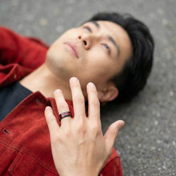 Laying on the ground, a man faces upwards with his hand stretched out. On his wedding finger is a black tungsten ring that has a groove of red running through the centre of the ring.
