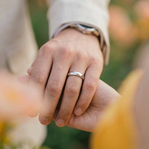 A zoomed in shot of a married couple holding hands. Blurred flowers frame the shot, with the mans ring prominent on his hand. It is a polished silver titanium ring.