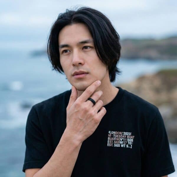 With a stunning beach scene in the background, a man stands with his hand to his chin. He wears a black silicone ring.
