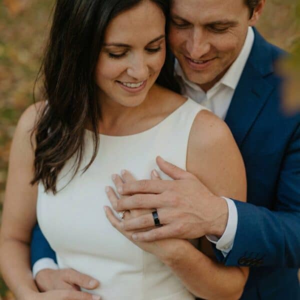 The newly married couple stand together with their hands joined. The man is wearing a flat style minimalist black tungsten ring that has a polished finish.