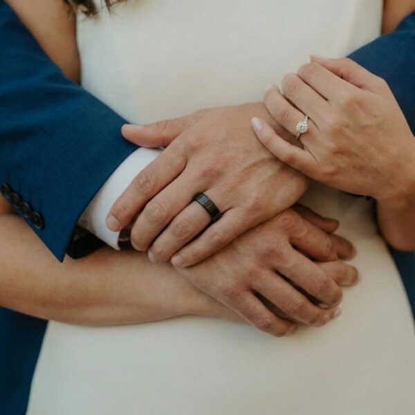 The bride and groom embrace while having their photo taken. This shot of the hands shows the couples wedding bands. The bride is wearing a diamond studded ring while the man is wearing an all black tungsten ring. This flat minimalist style ring has a polished finish.
