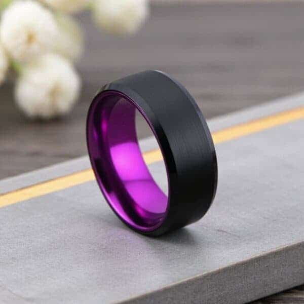 With flowers in the background, a ring stands upright on a grey and gold block. The ring is black and purple and is made with tungsten.