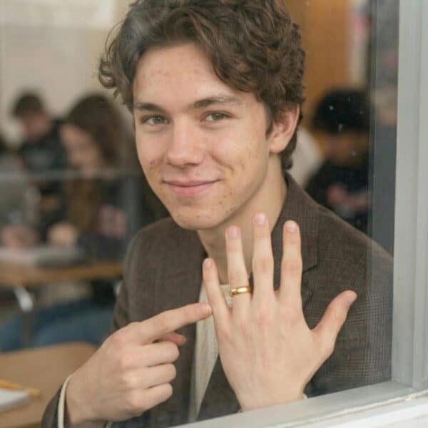 Smiling, a man holds up his hand and points to his gold titanium wedding band. With a polished finish.