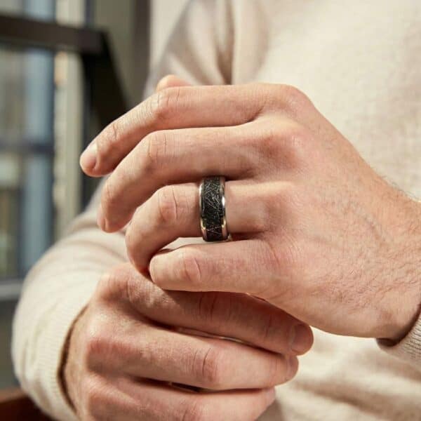 A close up of a man's hands. He is wearing a silver tungsten ring that features a distinctive black faux meteorite centre.