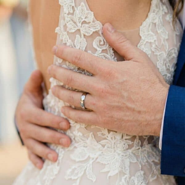 The couple come together for an embrace at the wedding ceremony. The groom holds his newly wedded wife. He is wearing a polished silver tungsten ring that is thin in style.