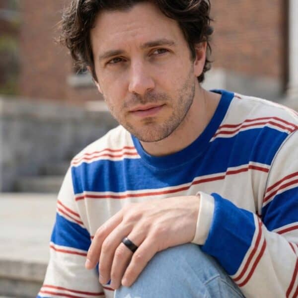 Looking into the camera, a man sits out with a wall and buildings behind him. Wearing a red, white and blue top with jeans, he is wearing a black ring on his finger. The ring is made with ceramic and has a brushed finish.