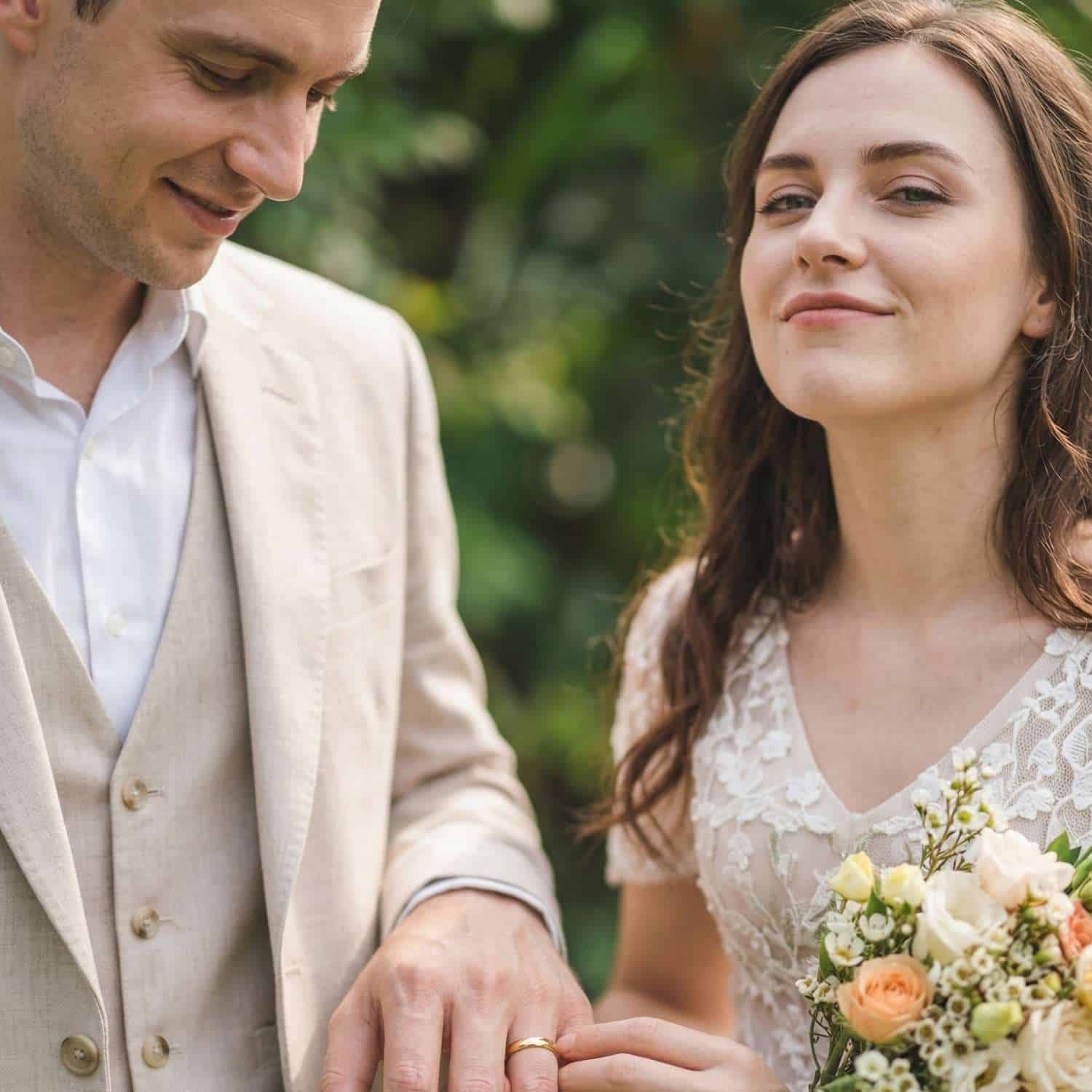 The newly married couple hold hands. The bride is wearing a white wedding gown and is holding white and light orange flowers. The man is dressed in a white shirt and a cream coloured suit. He is wearing a slim style gold tungsten wedding ring.