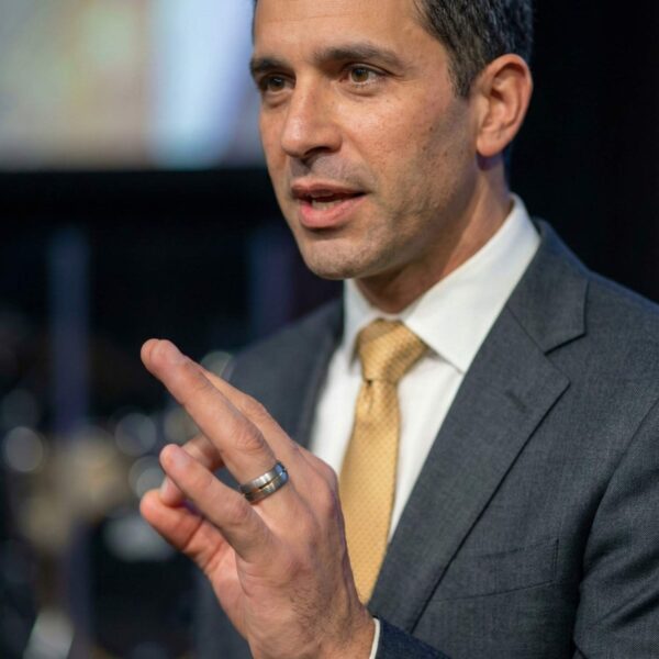 Speaking at a conference, the man is wearing a suit and tie. On his finger he is wearing a silver tungsten ring that features a groove of gold.