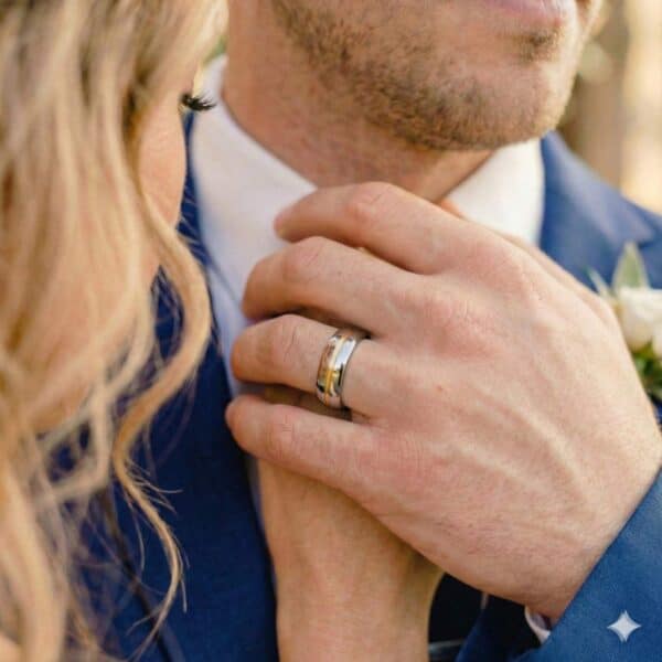 The newly married couple stand facing each other and holding hands. On the mans hand, he is wearing a silver tungsten ring that has a groove of gold running through its centre.