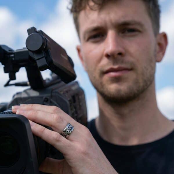 The camera operator takes a break from filming. Holding his equipment, he wears a silver and black ring on his finger. Made with tungsten, it features a cogwheel design.