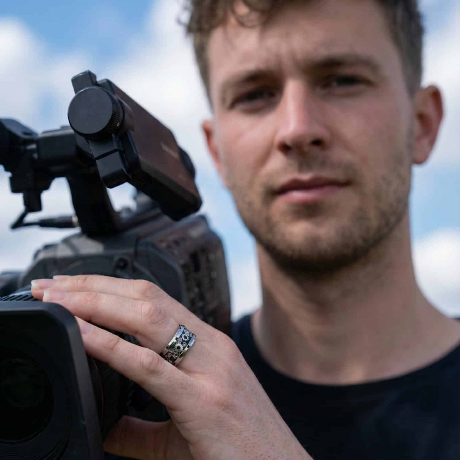 The camera operator takes a break from filming. Holding his equipment, he wears a silver and black ring on his finger. Made with tungsten, it features a cogwheel design.