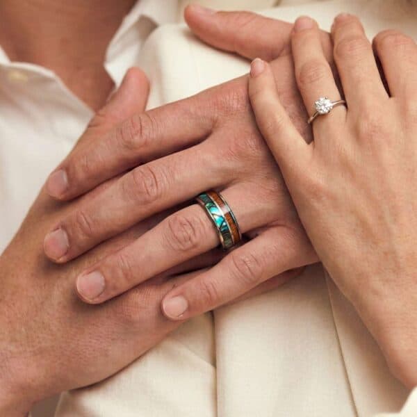A couple join hands, showing that they are both wearing wedding rings. The woman has a diamond ring while the man wears a tungsten ring that has inlays of natural wood and abalone shell.