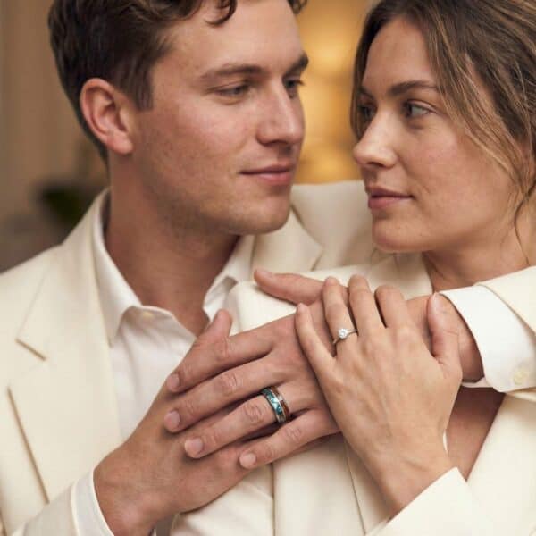 A couple gaze lovingly into each other’s eyes. Their hands are joined together. The woman is wearing a diamond ring, and the man has a silver tungsten ring that has inlays of natural wood and abalone shell.