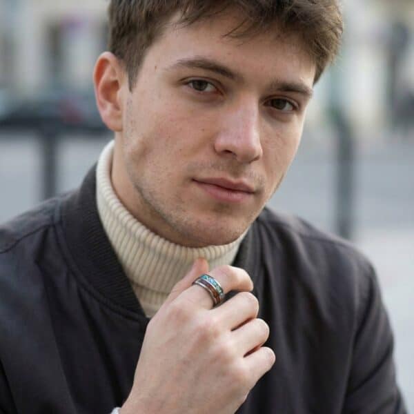 A man looks into the camera. Wearing a blazer and polo neck, he has his hand in front of his chest. On his hand, he wears a silver tungsten ring that features inlays of abalone shell and natural wood.