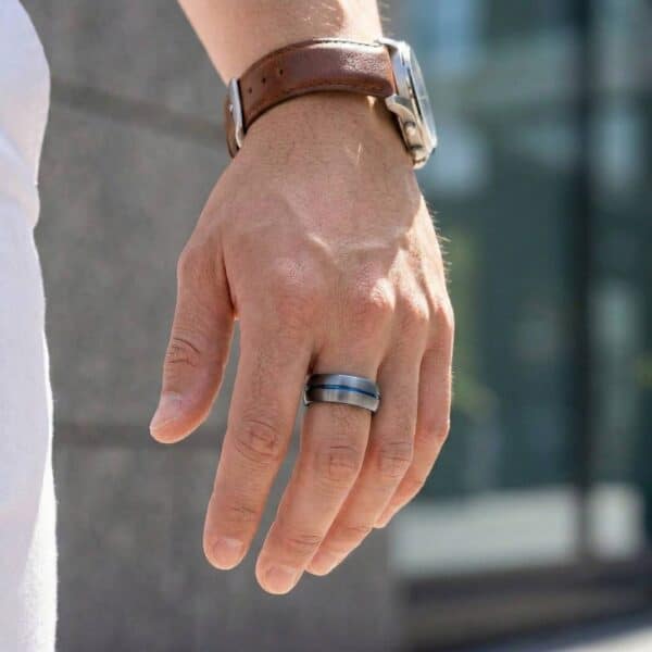 A close up image of a mans hand. One the middle finger, the man wears a ring. It is a gunmetal grey tungsten ring that has a groove of blue running through its centre.