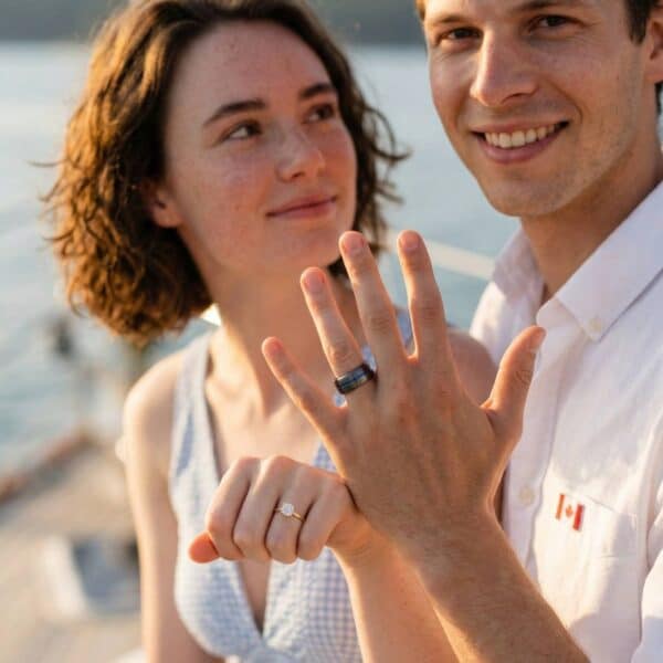 Just engaged, the happy couple raise their hands to show their new engagement ring. The woman has a gold engagement ring with a diamond. The man has a gunmetal grey ring with a vivid groove of blue, just off centre of the ring.
