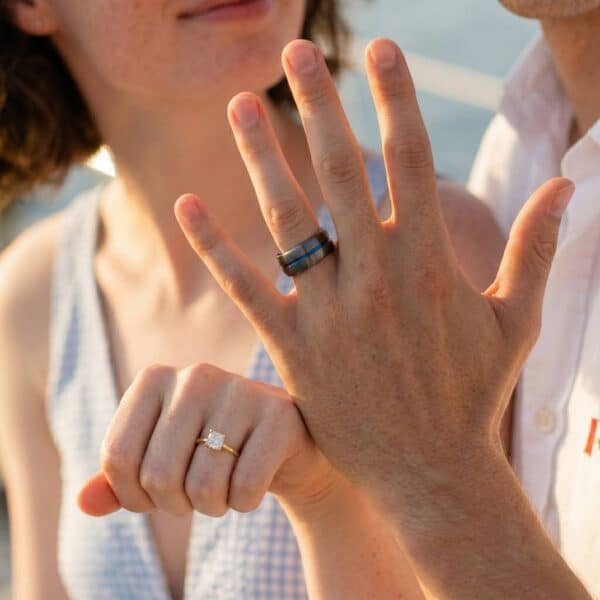 Holding up their hands to show their rings, the couple are delighted to show their rings. The woman has a diamond gold engagement band and the man has a gunmetal grey tungsten ring. It has a strip of blue running through the ring.