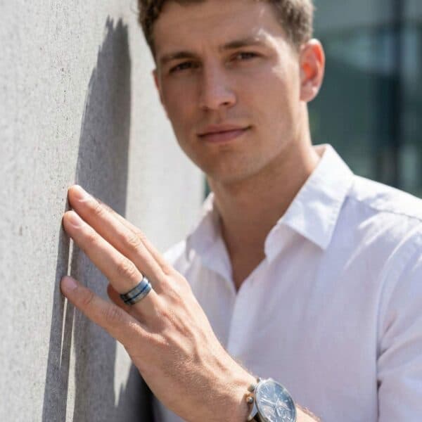 Leaning against a wall, a man wears a smart white shirt. On his hand he has a gunmetal grey tungsten ring that has a vivid strip of blue running just off centre of the ring.