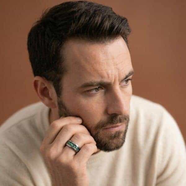 A man with a beard sits facing the camera but looking slightly away from the lens. With a thoughtful expression, he has his hand to his face. On his wedding finger he wears a silver tungsten ring that features an abalone shell inlay.