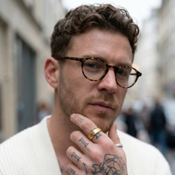 Looking into the camera, a man wears glasses and has his hand held to his chin in a thoughtful way. On his index finger, he is wearing a gold tungsten ring. It has a brushed centre section and polished gold edges.