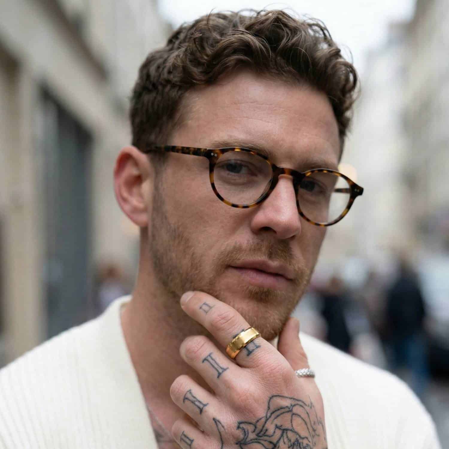 Looking into the camera, a man wears glasses and has his hand held to his chin in a thoughtful way. On his index finger, he is wearing a gold tungsten ring. It has a brushed centre section and polished gold edges.