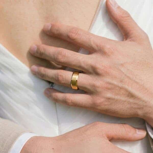 A close up image of a bridge and groom as they hold each other. You can see the back of the bride while the man has his arms around her. You can see his hands in detail. He is wearing a gold tungsten ring. It has a brushed surface and polished bevelled edges.