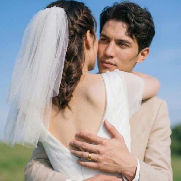 On a bright summers day, a couple embrace on their wedding day. The bride, wearing her white wedding gown has her back to the camera. You can see the mans face. He has his arms around his partner. He is wearing a gold tungsten ring on his hand. It has a brushed flat surface and polished bevelled edges.
