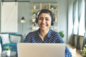 Head shot cheerful smiling pretty hindu ethnic girl sitting at table with computer, wearing headphones with mic, looking at camera