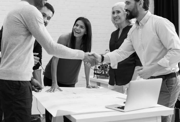 Individuals standing at a table during a meeting with two men shaking hands
